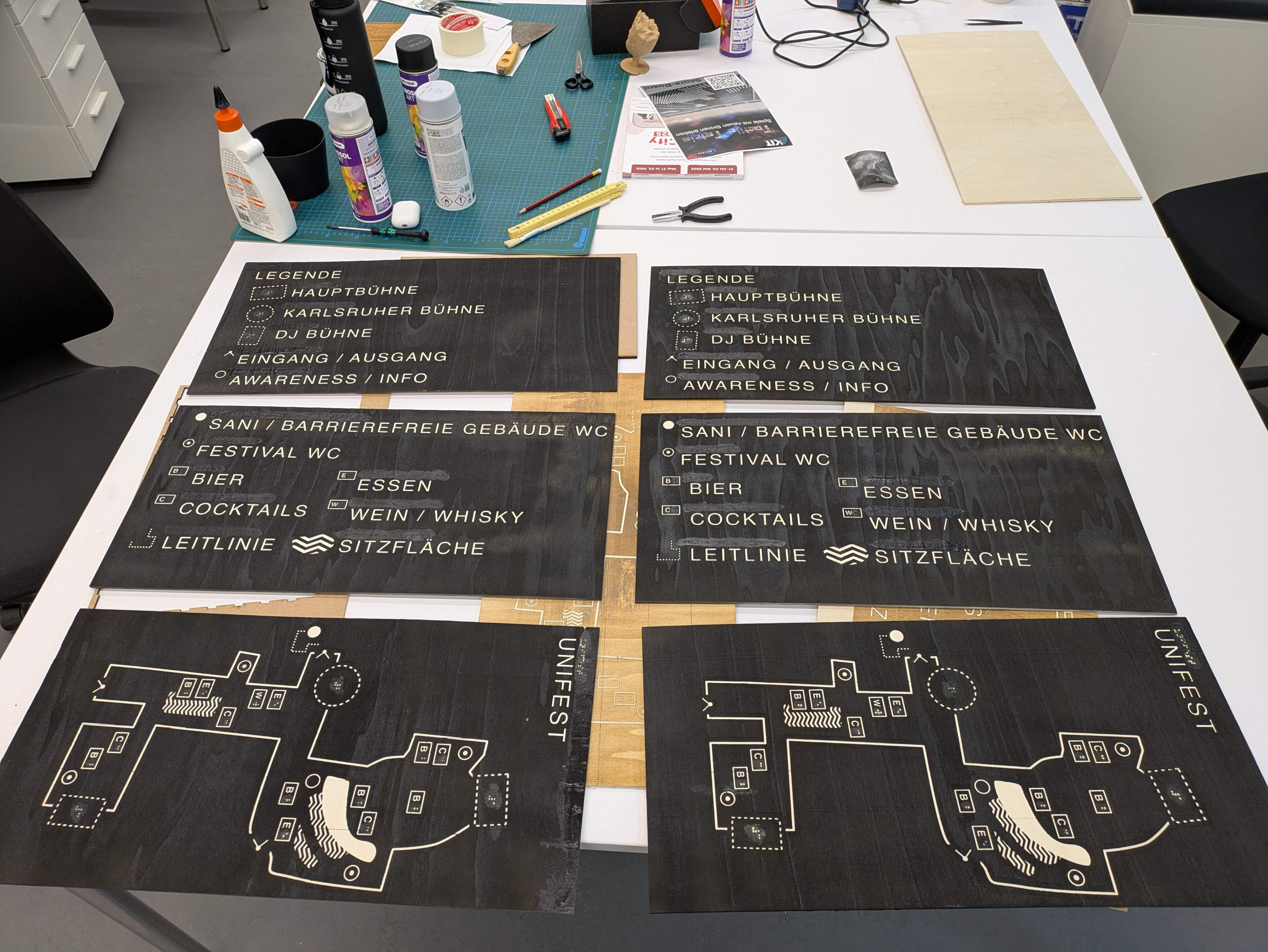 Six black wooden panels are arranged in a 2 x 3 pattern on a white table.
All raised elements are unvarnished.
The black varnished background allows the grain of the wood to show through.
In some places, instead of wood grain, there is an surface smooth strip of wood glue, which is uniformly black.
Above the panels, there are many objects scattered on the table. Among them: a pencil, a craft knife, tweezers, wood glue, spray cans, and a wooden 3D model of Groot's head from the Marvel movies.