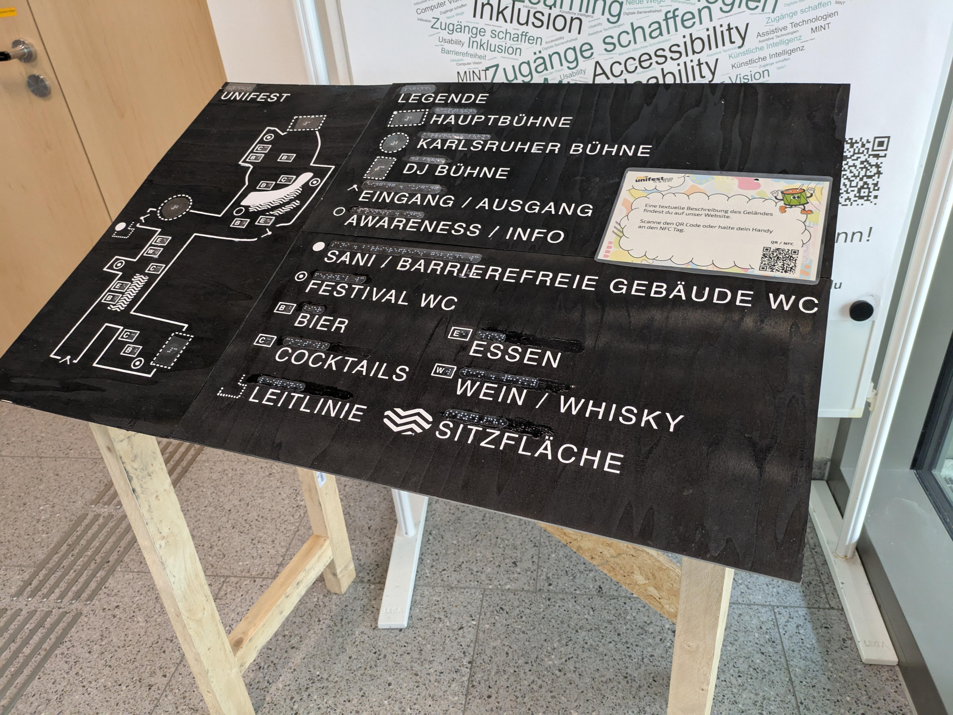 A close-up of the same tactile site plan.
This time without people and indoors.
The floor consists of light-colored tiles with dark speckles.
In the background, parts of a floor guidance system, a door, and a display stand with a poster are visible.
A laminated DIN A5 sign has been added to the map in the middle, adjacent to the right edge.
It is labeled (in German) “A textual description of the terrain can be found on our website. Scan the QR code or hold your cell phone to the NFC tag.”
The text is in black and has been embossed with Braille.
In addition to the QR code at the bottom right, there are other graphic embellishments such as the lettering “Unifest 2025” at the top left and a drawing of a drum with a face, arms, and legs happily whistling at the top right.