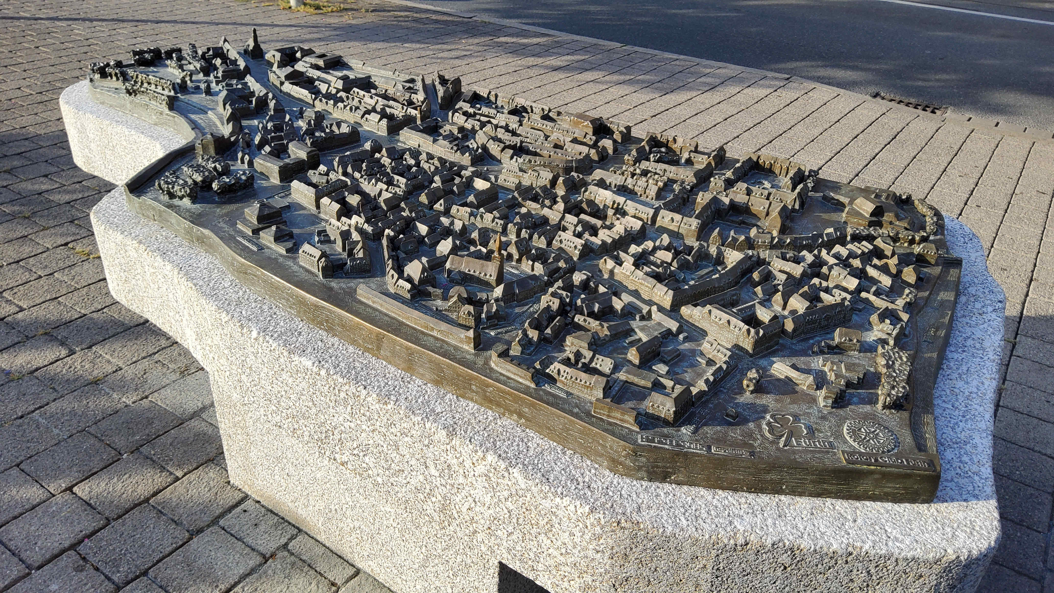 A photo taken at an angle of a tactile city model of Fürth, made of bronze. It is visible on a granite base at approximately waist height. The buildings and all elements are cast from a single mold. They feature a level of detail that includes individual church towers and skylights of residential buildings. The names of the streets are written in standard lettering with profile and Braille.