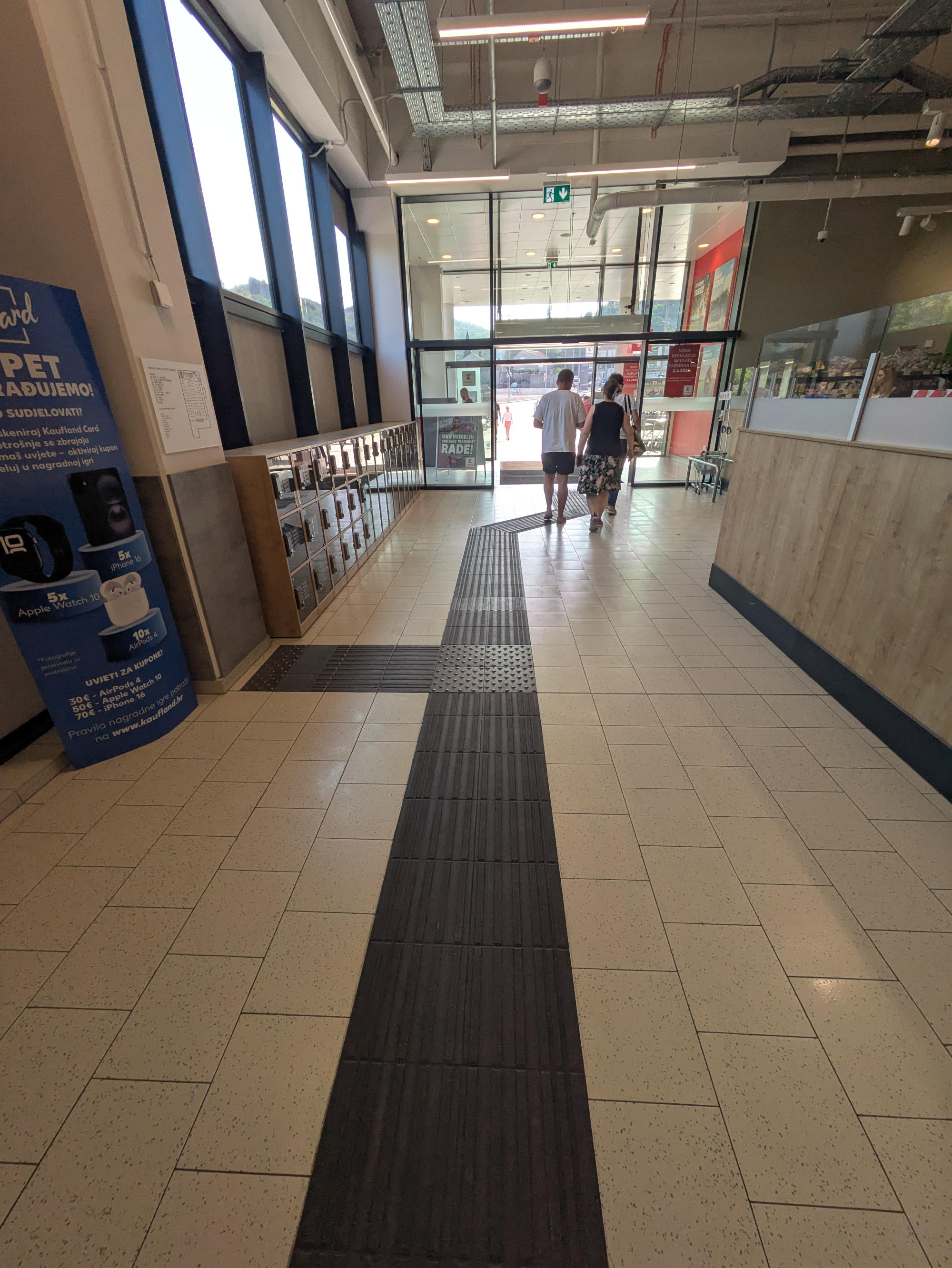 A photograph taken from inside the supermarket, facing the entrance doors.
On the left, the tactile map is visible on the wall; on the right, a room divider separates the entrance area from the sales area behind it.
In the center, three people in summer casual wear are walking toward the exit with their backs to the camera.
The floor has light-colored tiles. A black tactile floor guidance system runs down the center.
The entrance is slightly offset from the path. The floor guidance system follows this offset with two 45-degree bends without interruption.
Behind it is an attention field with an orthogonal branch leading to the tactile map.
