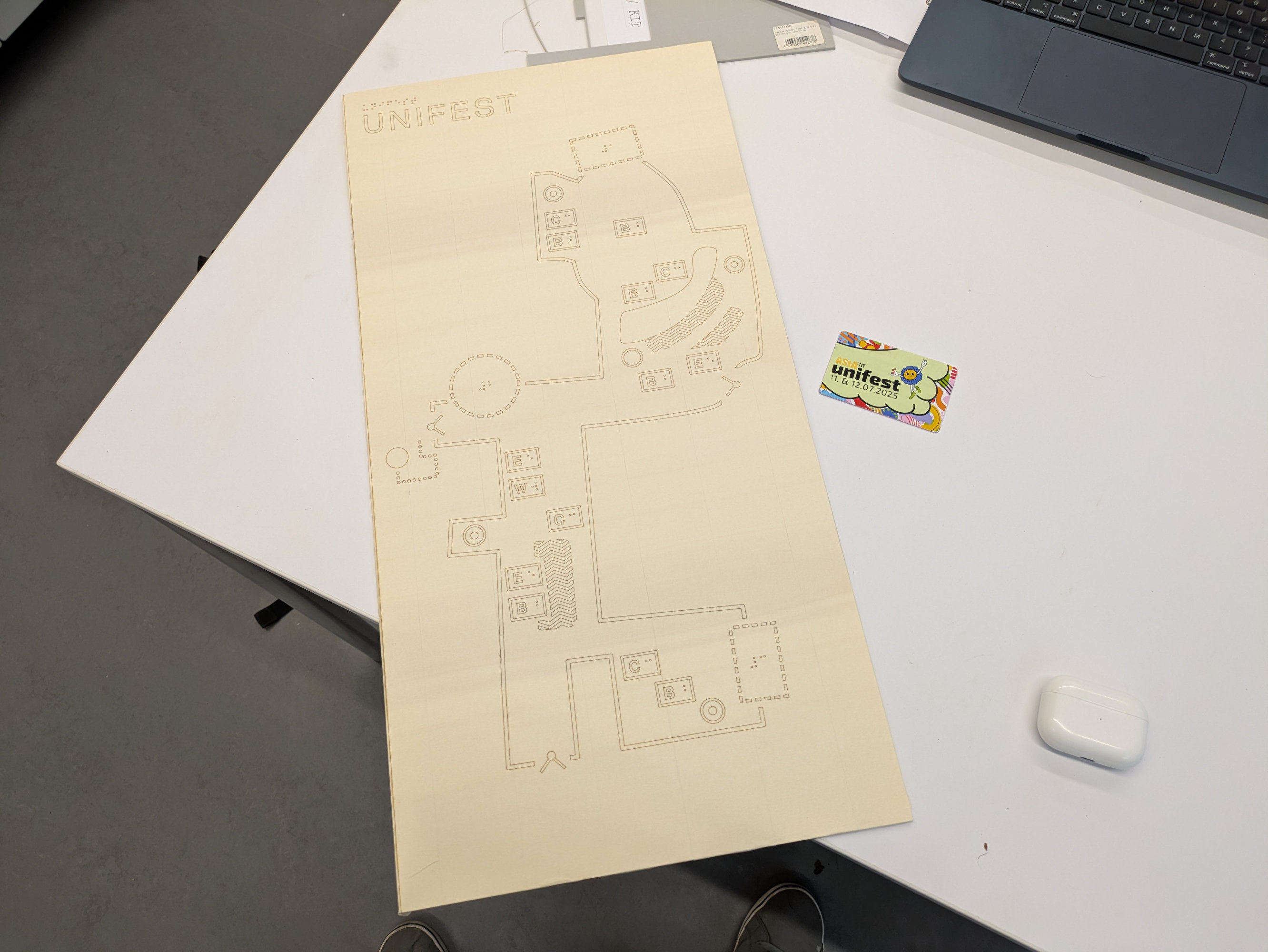 A photo of one of the wooden panels.
It is completely covered with masking tape.
The contours of the map are cut along the tape.
The panel is lying on a white table.
Next to it is a student ID card with a sticker labeled “Unifest” and a drawing of a smiling sunflower.