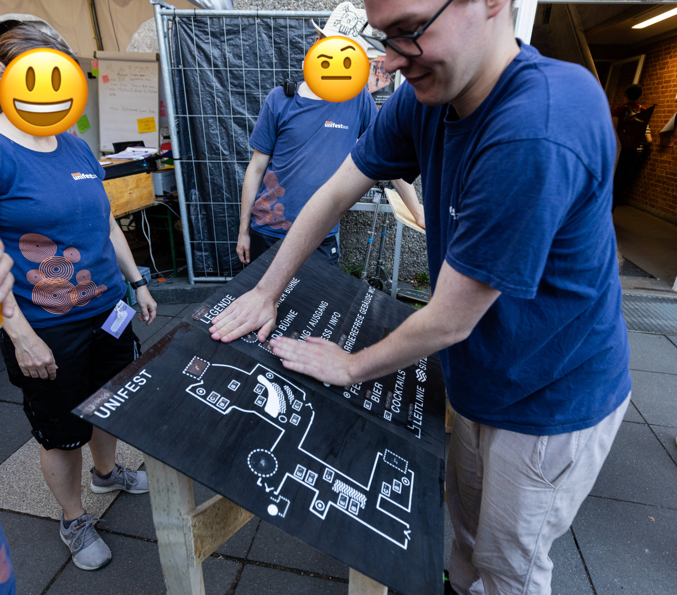 A photo of me pressing down on the three finished wooden panels with both palms on a wooden table.
The table is waist-high and angled at 30 degrees.
The table consists of untreated but sanded, sawn frame wood segments and a cut-to-size chipboard panel, which connects two legs crosswise for stabilization.
There are two other people in the picture next to me.
We are all wearing the same dark blue T-shirt with “Unifest” printed on the chest.
The faces of the other two have been edited with a 😃 emoji and a 🤨 emoji.
The picture is taken outdoors.
In the background are a front desk, a flipboard with notes and Post-its, and a construction fence.