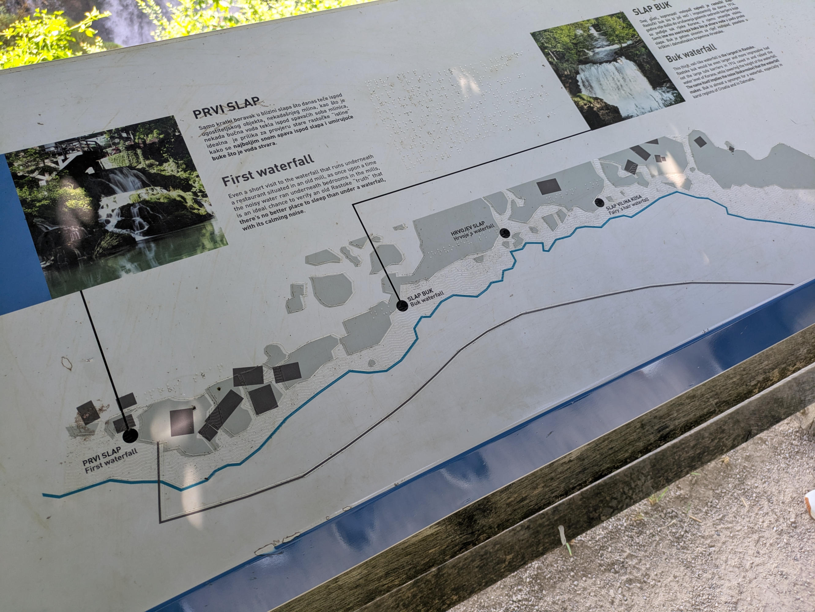 A photo taken at an angle of a wide information board approximately 50 cm high and 2 meters wide. However, the board protrudes from the side of the image. The lower half shows a schematic visual representation of a river course with adjoining banks. Fixed objects on the riverbank are represented tactilely with a raised line, while water is represented by a wavy line that can be felt. The tactile elements themselves are applied transparently to the color print. Some visual elements have no tactile counterpart. These are not only omitted, but also represent a different meaning with the tactile pattern.
The upper half contains images and information texts about nearby waterfalls in Croatian and English. Next to the print text is a text written in Braille, which could reflect the length of one of the languages. The Braille is also transparent. The print has a font size of approx. 0.5 cm.
Black reference lines lead from the text blocks to the lower half of the map.
The lines have no tactile equivalent.
