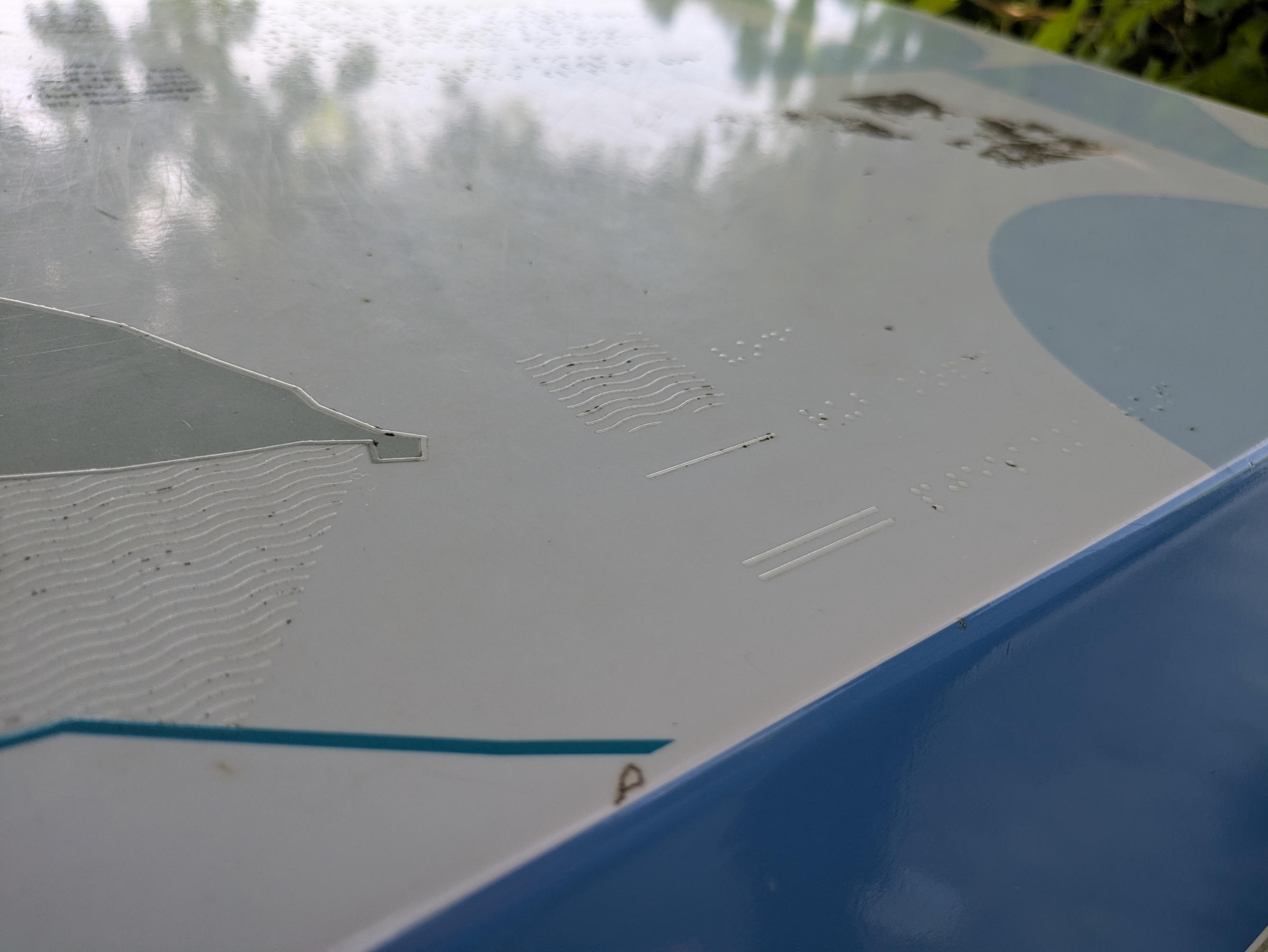 A low angle image of the same information board, more detailed. Only a small section of approximately 10 x 10 cm is shown.
There are three lines. Visually, these are white backgrounds. Tactilely, there is always a symbol and a word in Braille.
In line one: a tactile wave pattern, line two: a horizontal line, line three: two parallel horizontal lines.
Due to the flat angle, the sun reflects off the raised elements, making them easier to recognize.
The legend is not marked as such and is located on the board without any recognizable nearby clues.