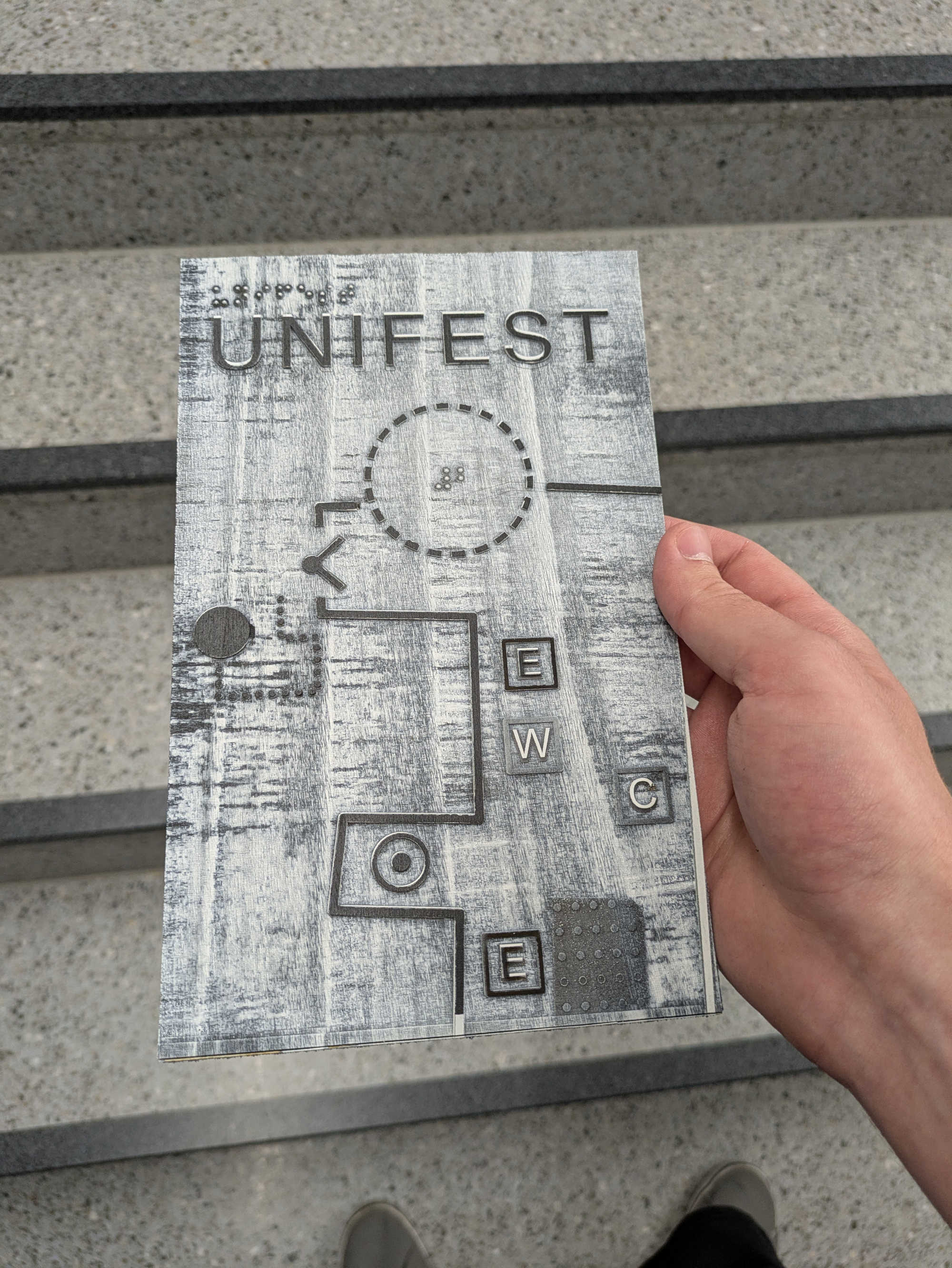 A photograph of a thin wooden board measuring 10 x 20 cm, held by a hand at the edge.
It shows a section of the tactile map design of Unifest.
The background is painted white. The paint is uneven; in some places, it does not seem to have adhered at all.
Raised elements are painted black.
In some places, the edges are not cleanly black, but white.